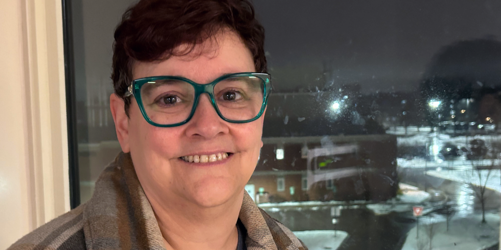 a person with short brown hair, wearing jade-colored glasses and a brown plaid jacket stands in front of a dark window