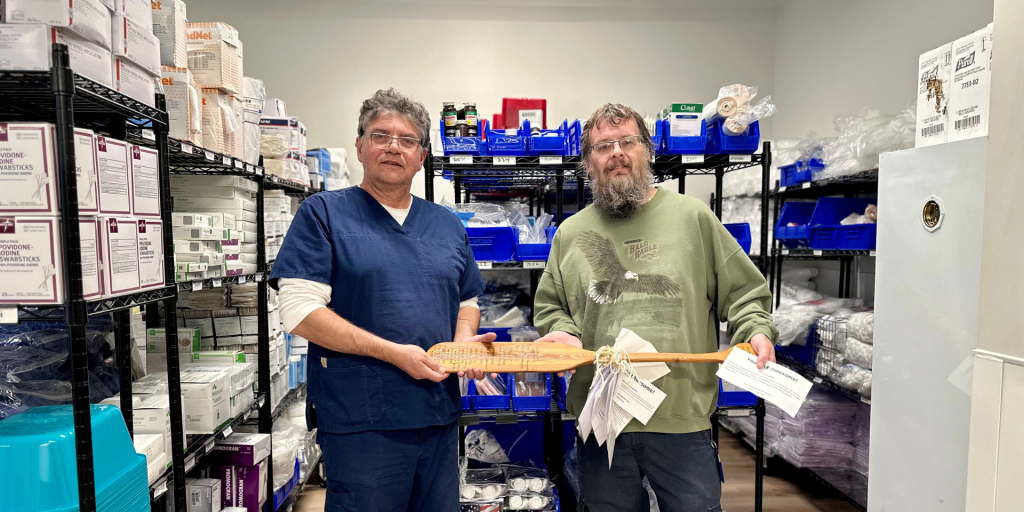 Two men, one in a blue scrub and the other in a green shirt, standing in a storage room holding a paddle.
