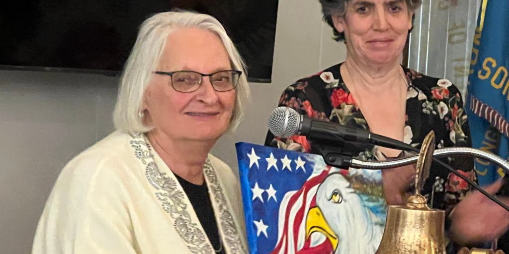 Two women at a speaking event with one presenting an eagle painting while standing at a microphone.