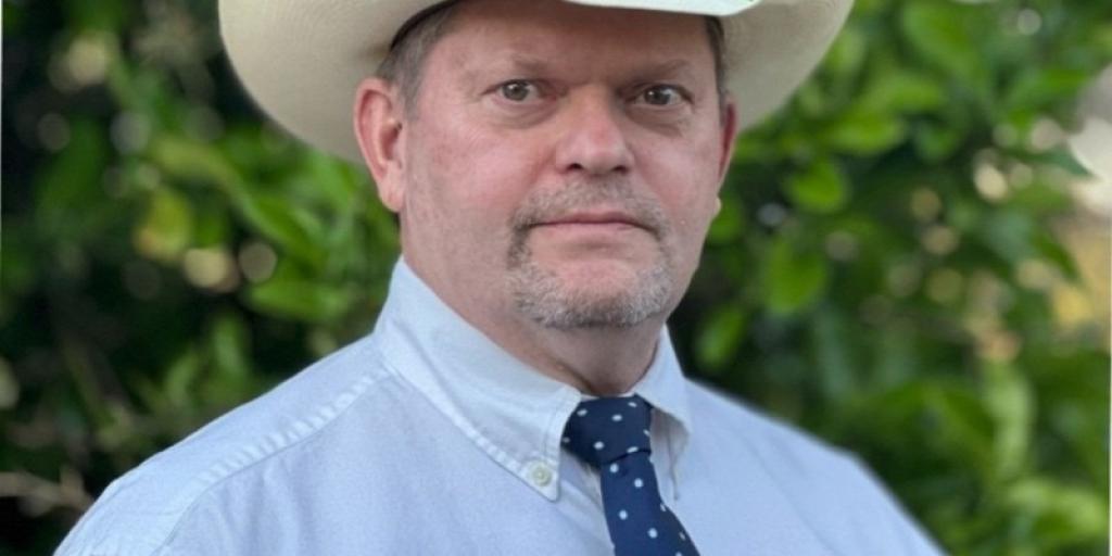 Man standing in a button up shirt and blue polka dot tie, wearing a cowboy hat.