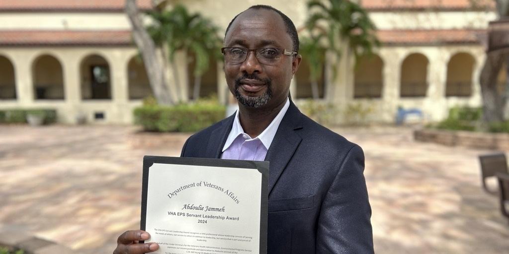 A man who is dressed in business casual attire stands outside on a sunny day, holding the award that he earned.