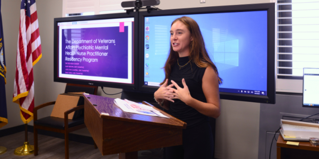 Young female nurse wearing a black sleeveless blouse speaks to the audience about her project while standing at the brown podium.