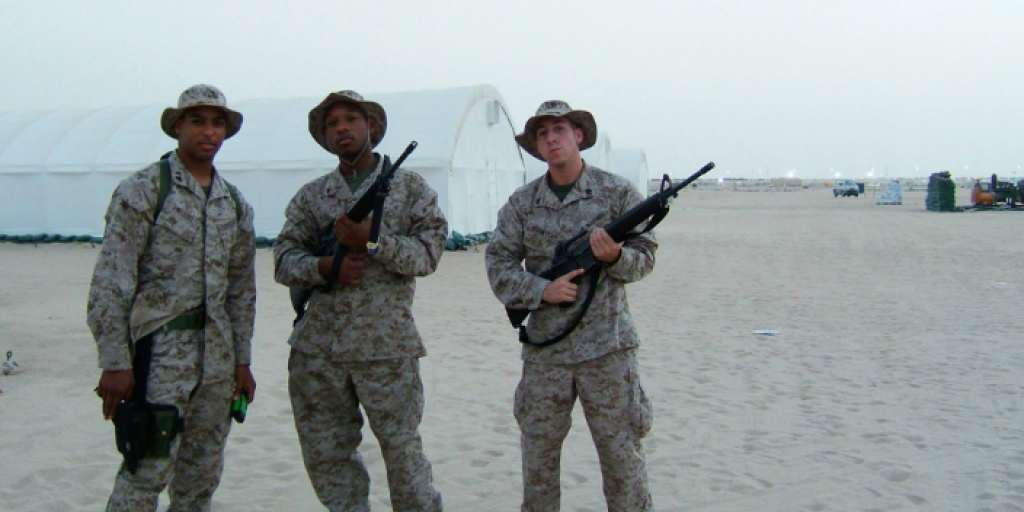 3 U.S. Marine Corps Veterans standing on base in Iraq with their guns facing the camera with sand and tents in the background.