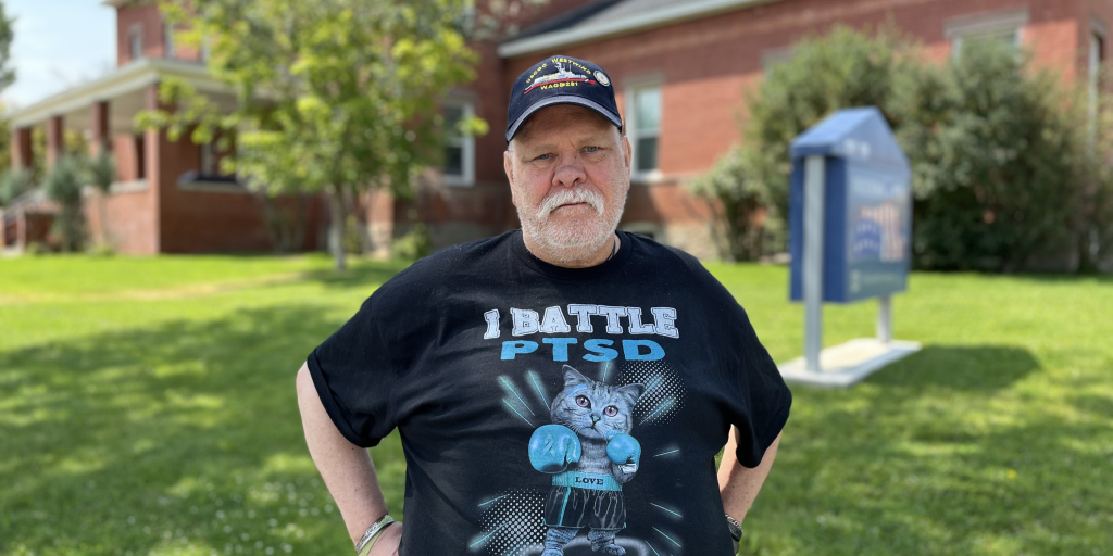 A man standing in front of a brick building, wearing a black baseball cap and a black t-shirt.