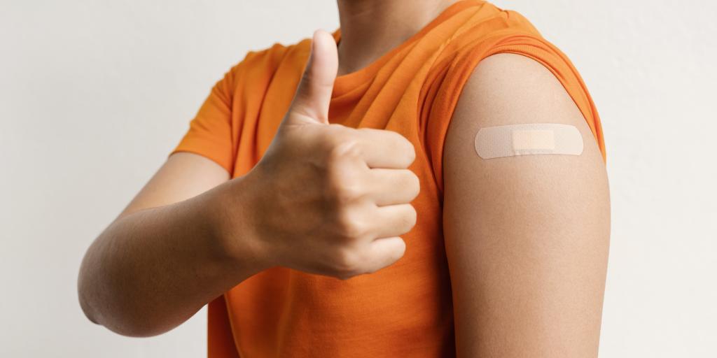 person in orange shirt with bandaid on shoulder and holding a thumbs up.