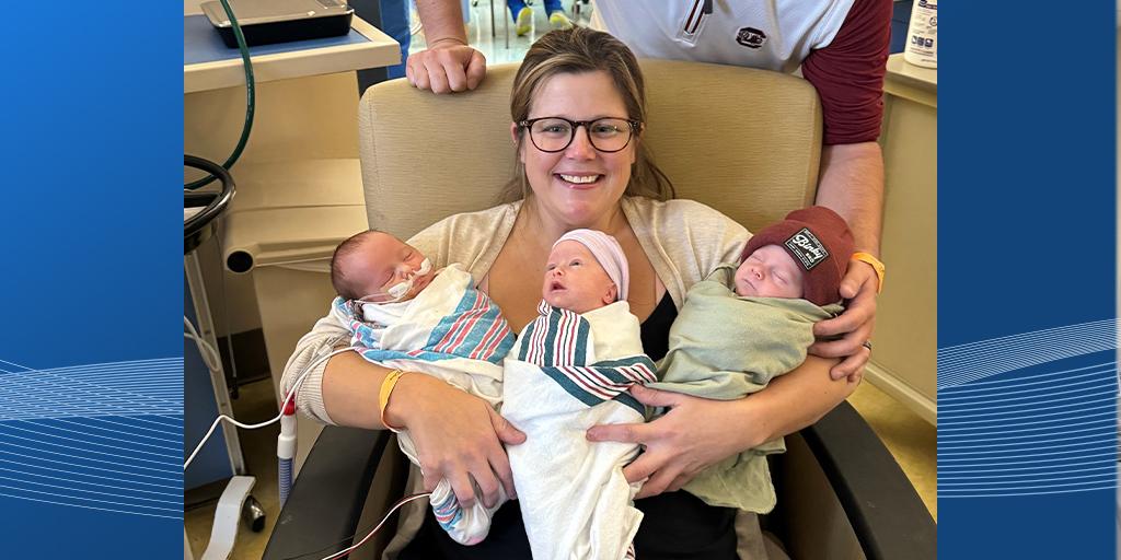 Dibble sits in a chair, smiling, while holding her triplets.