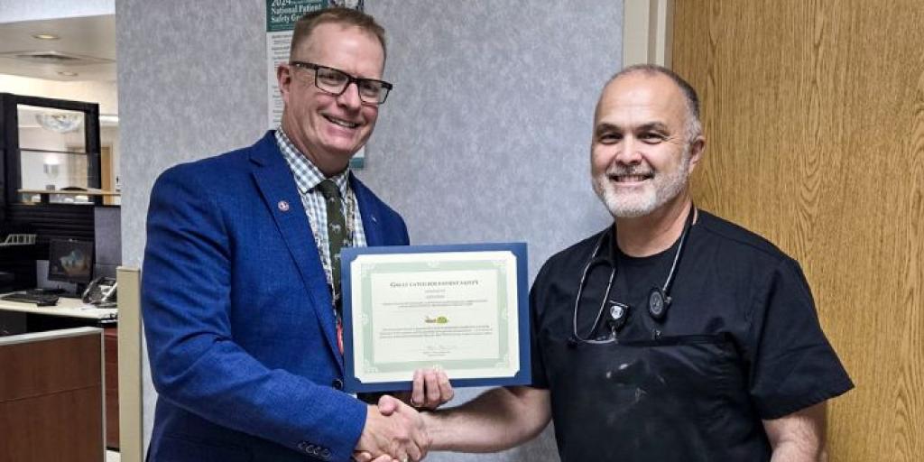 A man wearing a blue suit and a male nurse shake hands and smile while receiving a Great Catch Award certificate.
