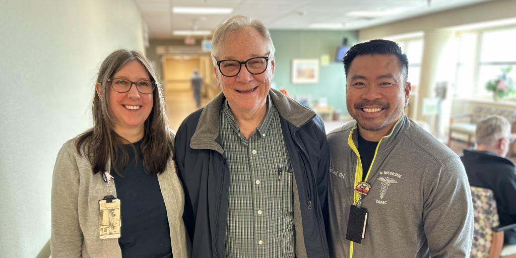 Man stands in the middle of a hospital hallway, wearing a checkered shirt, blue jacket and black glasses. On either side of him are his care team.
