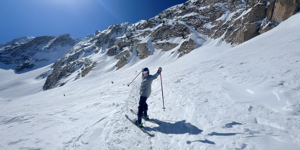 A person is skiing on a snowy mountain slope with a backdrop of snow-covered peaks and a clear blue sky.