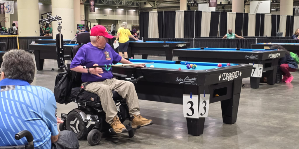 A man in a wheelchair engaged in a friendly pool game with several other players around him.