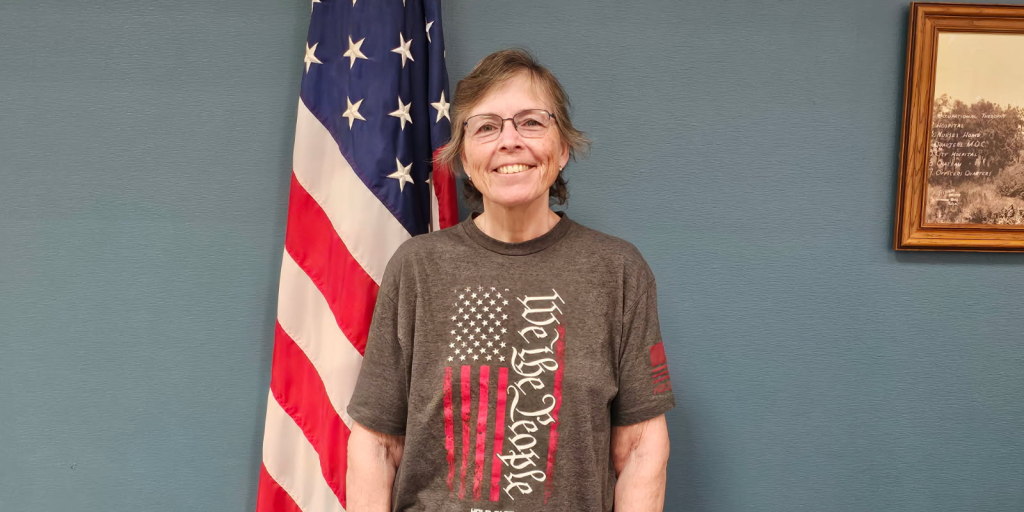A woman wearing a t-shirt featuring the American flag, standing confidently with a smile.