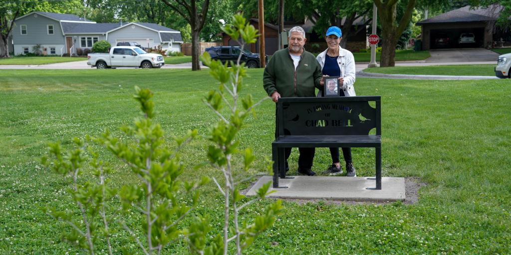 A man and a woman stand behind a black metal bench holding a framed photo. the bench is inscribed with "In Loving Memory of Chad Bell" 