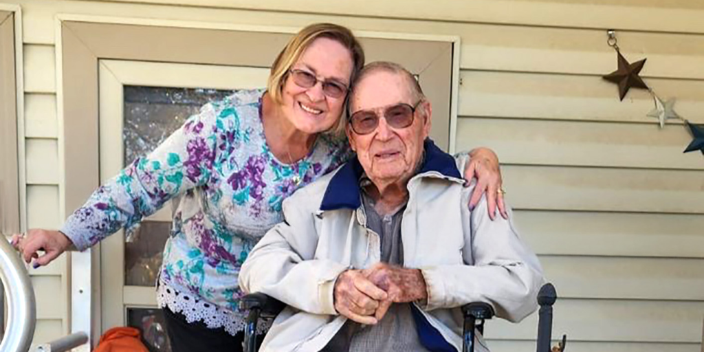 On a porch, Henry White and Carla Holuby sit together, surrounded by pumpkins, enjoying a peaceful autumn day.