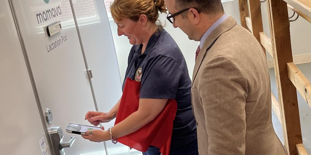 Caucasian woman in blue scrubs holding a cell phone as Caucasian man looks on. Both stand in front of the new Mamava lactation pod.