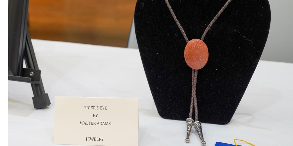 A glittering red bolo tie sits on a stand next to a blue ribbon