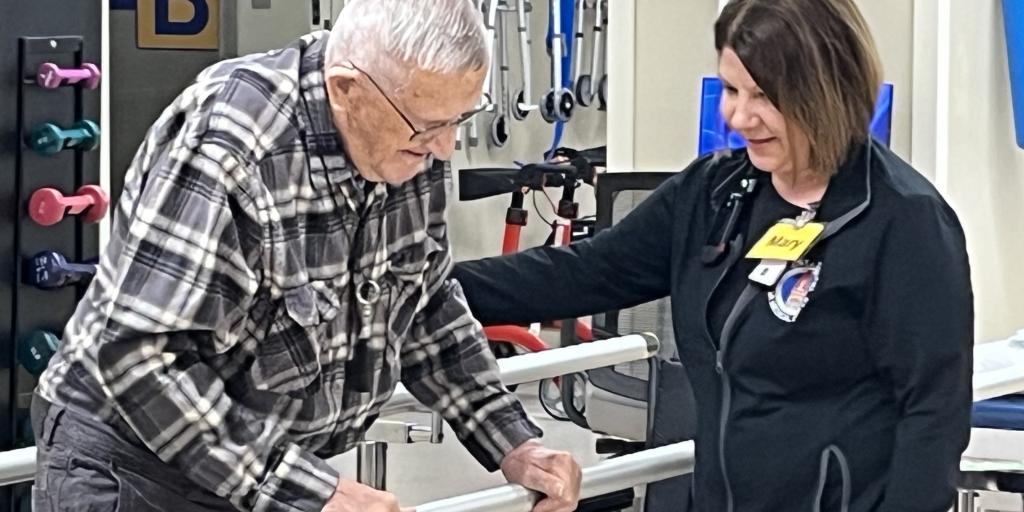 A man shuffles sideways, holding a bar, while a woman watches over him. 