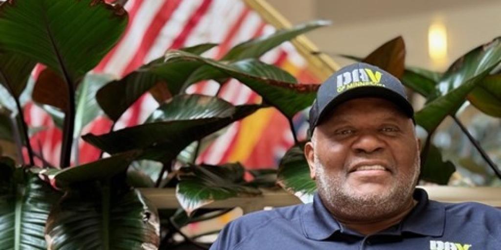 an man with dark skin wearing a DAV baseball hat and shirt sitting in front of plants with a flag in the background