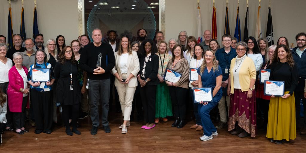 A large group of people posing in front of flags and a VA seal at the Spirit of George E. Wahlen Awards ceremony
