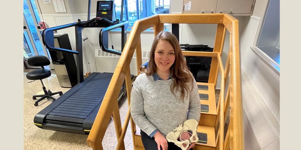 Female seated on therapeutic steps near a treadmill holding a 3D skeletal model of a pelvis