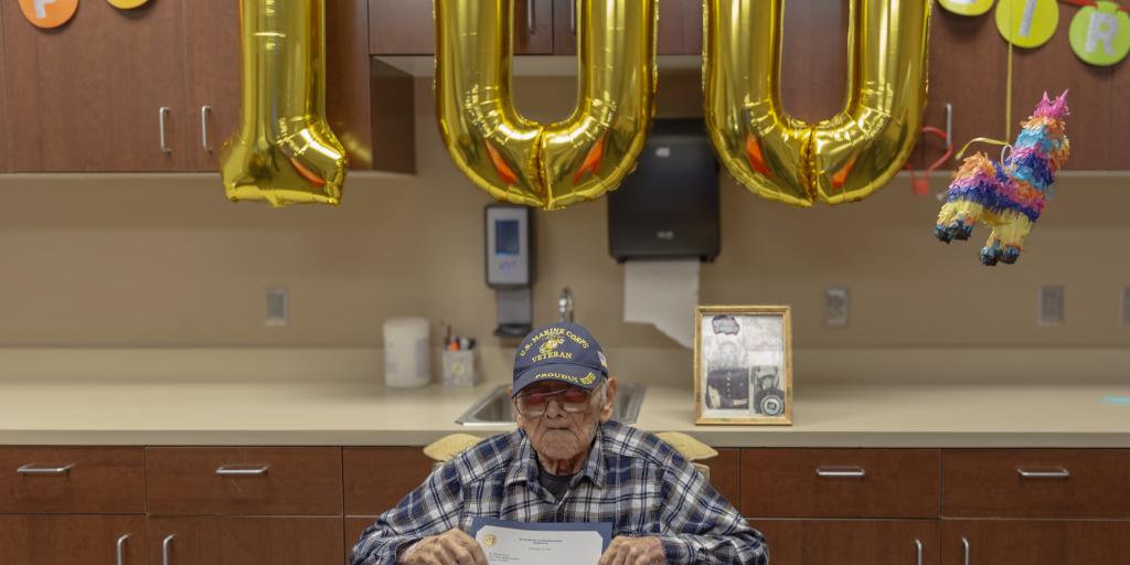 Veteran in wheelchair holds certificate in front of 100th birthday balloons.