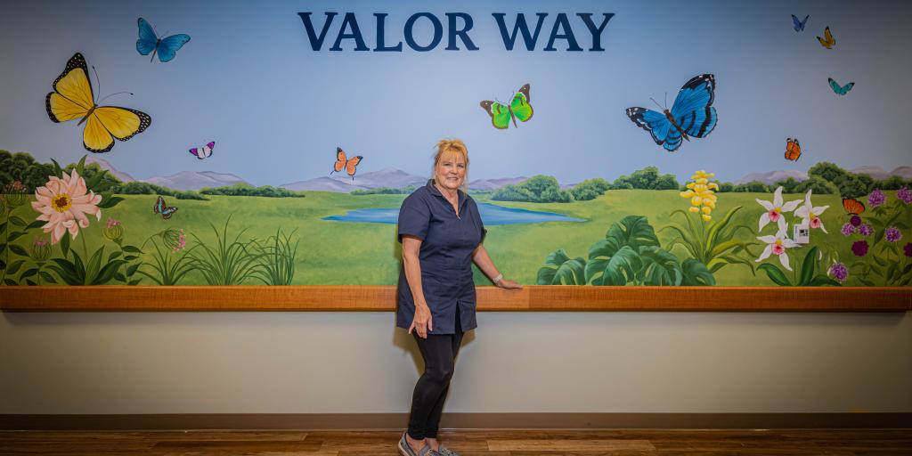Woman stands in front of mural with butterflies, flowers, and landscape.