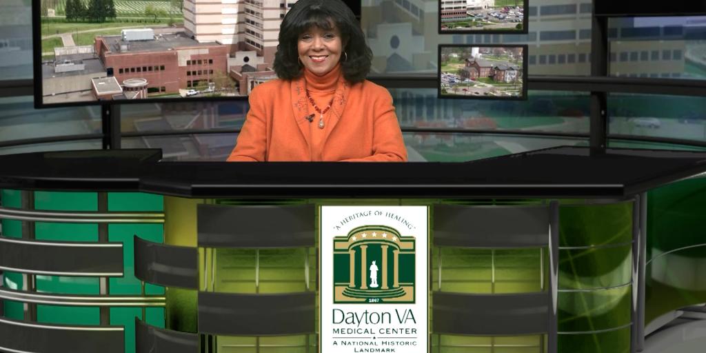 Woman sitting at a green and black news desk with a Dayton VA logo.