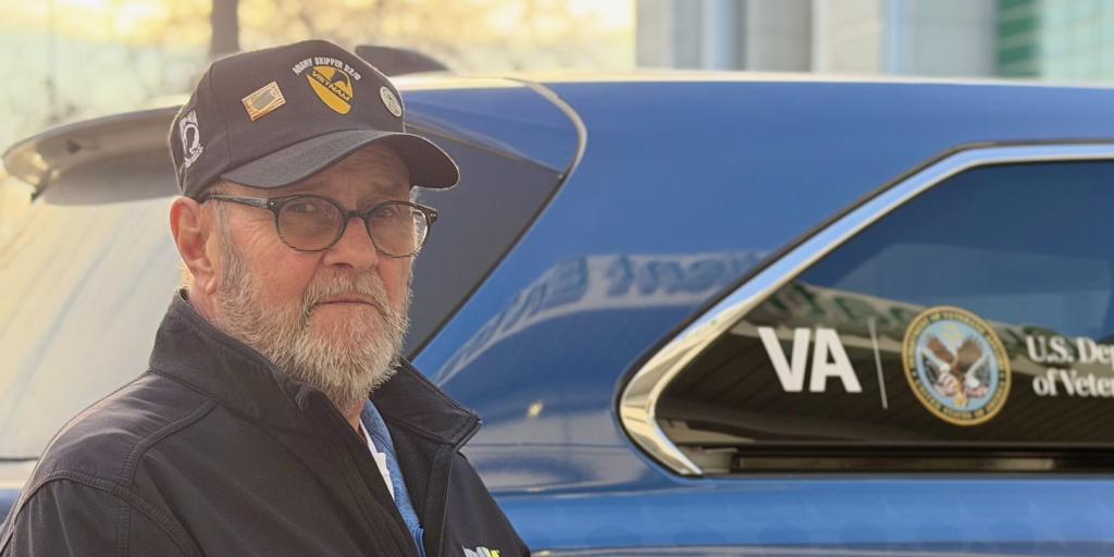 Man with cap next to blue VA Department vehicle.