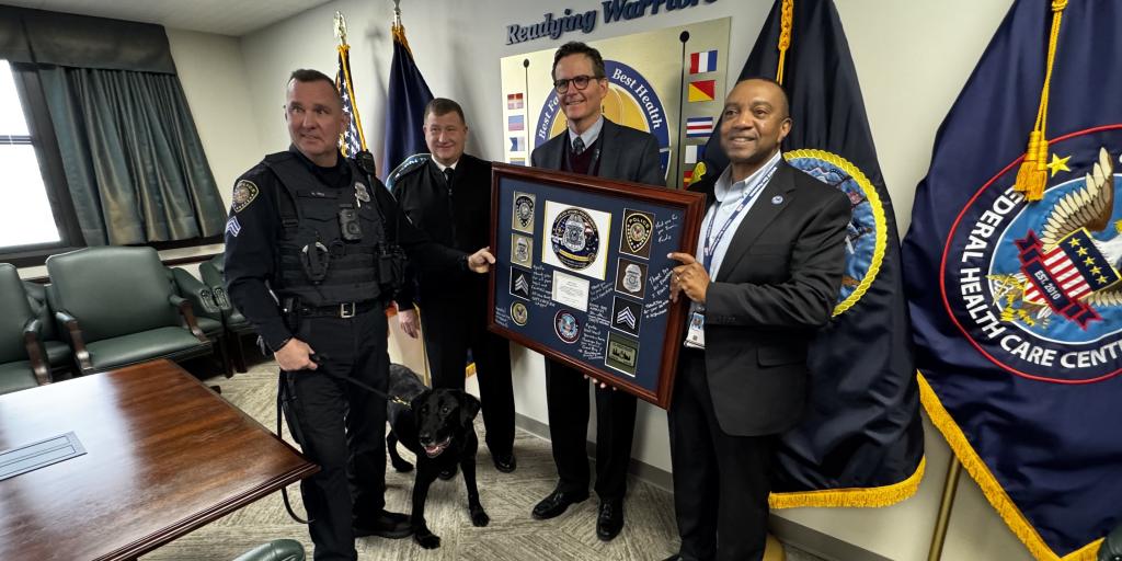 A police officer and three men in suits with dog stand in a room with flags, one holding a plaque.
