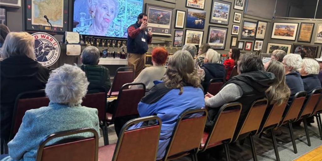 Man standing and speaking in front of a large screen, audience seated in chairs watching.