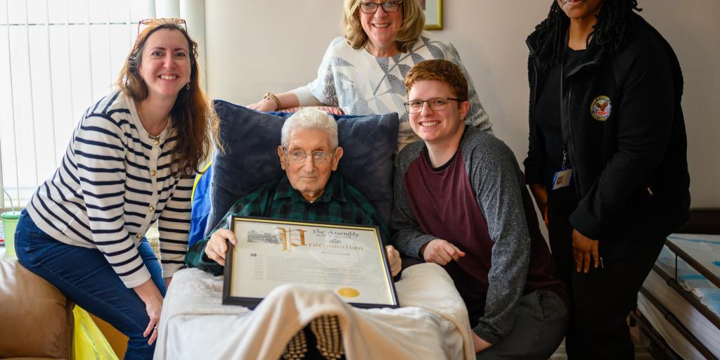 Five people in a living room, one is an elderly man in a chair with a plaque.