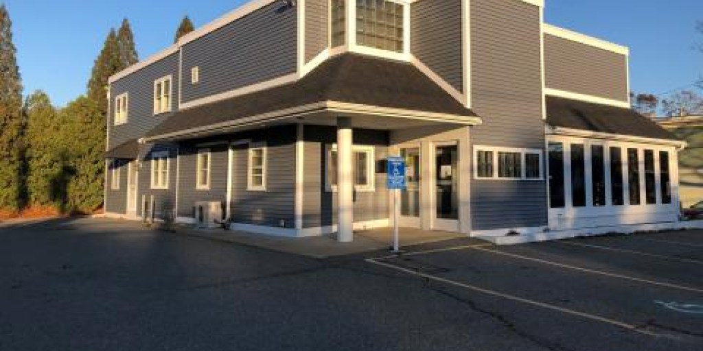 Gray building with black trim, multiple windows, and blue sign in front of parking lot.