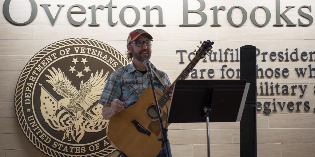 Man with beard and glasses holding guitar in front of Overton Brooks VA Medical Center sign.