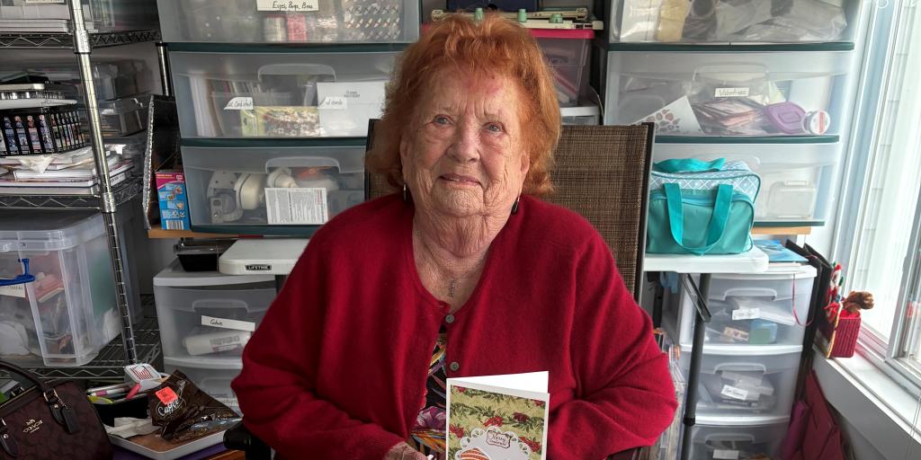 Elderly woman in red sweater reading a book, surrounded by storage boxes.