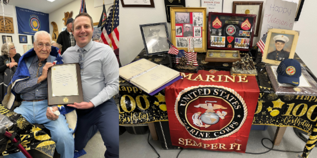 Man presents framed certificate to seated man in front of Marine Corps banner.