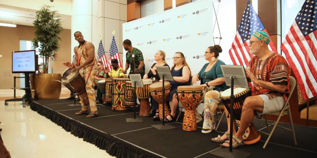 Diverse group of people performing on stage with drums and laptops, American flags in background.