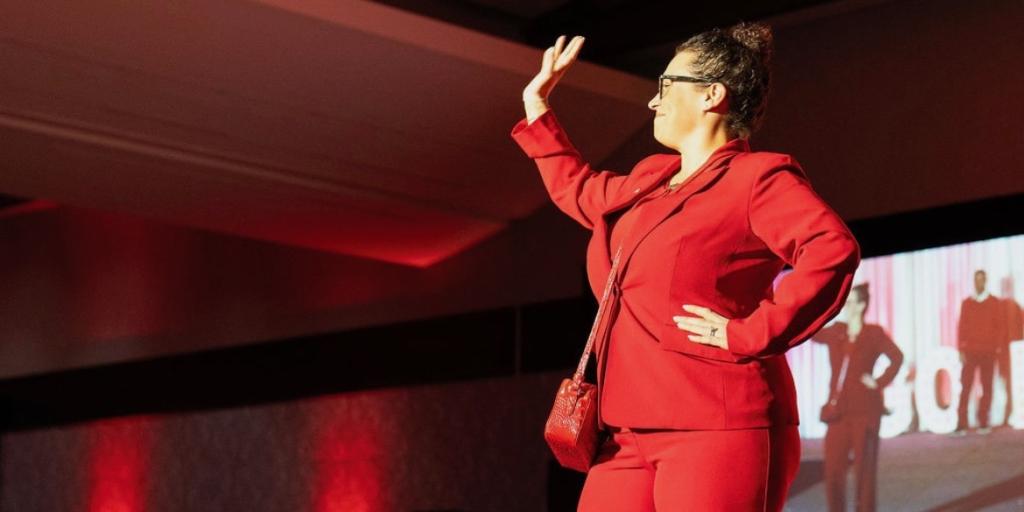 Woman in red suit standing on stage with arm raised, audience in background.