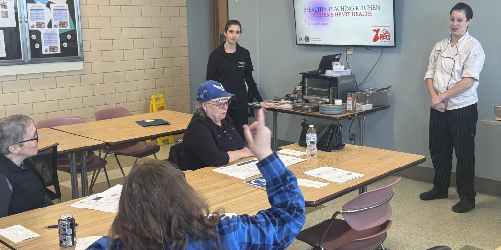 Three Women Veterans sit at tables in a classroom kitchen space listening to a VA Providence staff member who is presenting at the front of the room. Educational handouts and menus are spread across the tables during the Healthy Teaching Kitchen session.