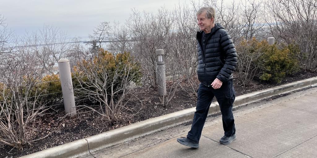 Man in black puffer jacket and jeans walks on sidewalk near shrubs and water view.