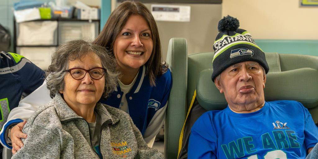 A smiling woman and man sit in chairs with a young woman standing behind them.