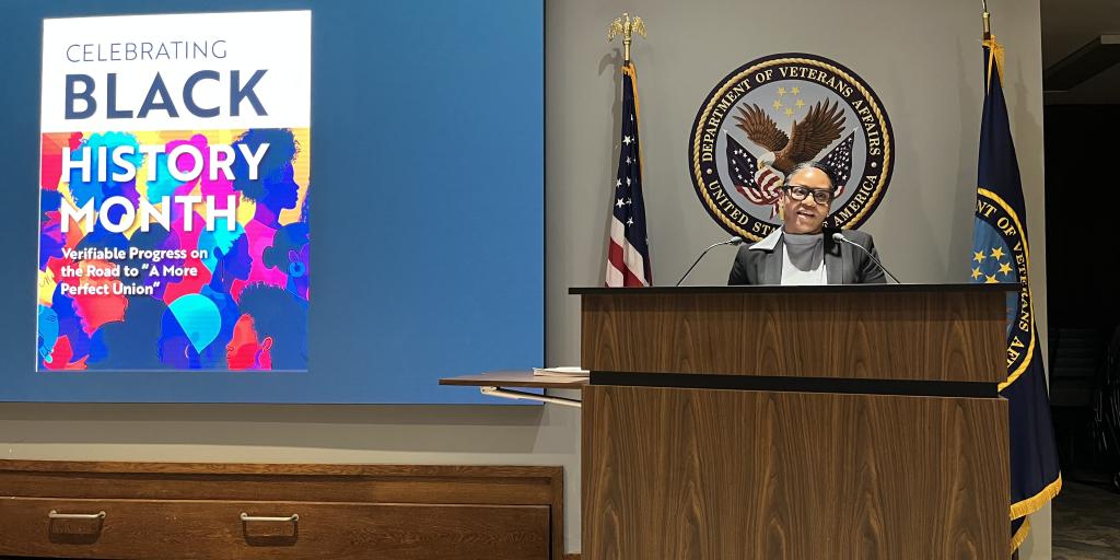 Speaker at podium with "Celebrating Black History Month" poster in background.