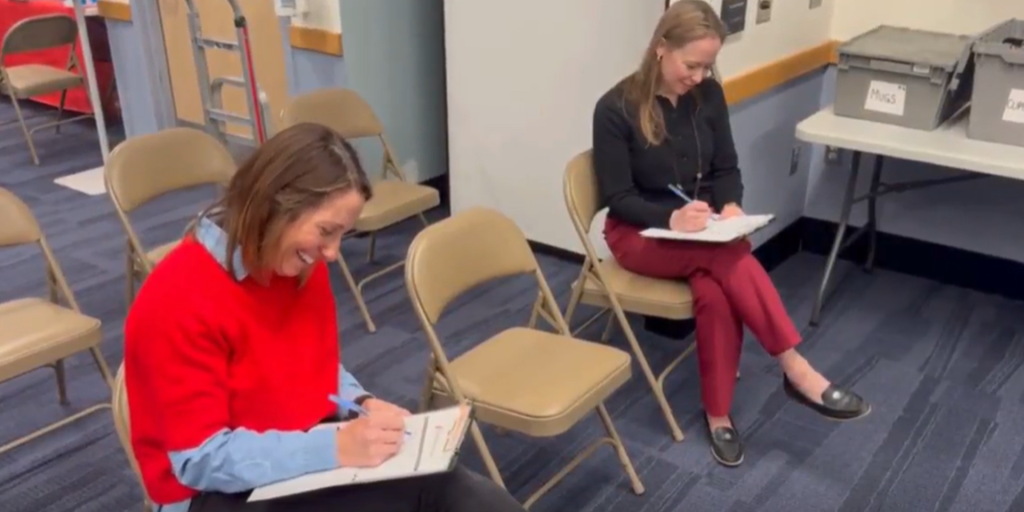 Two women in chairs write on paper with pens.