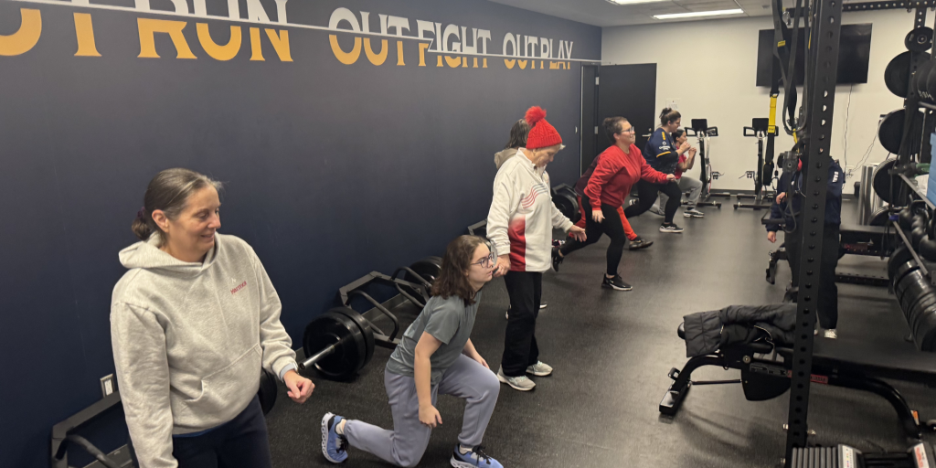 Six people in a gym. One person is doing lunges.