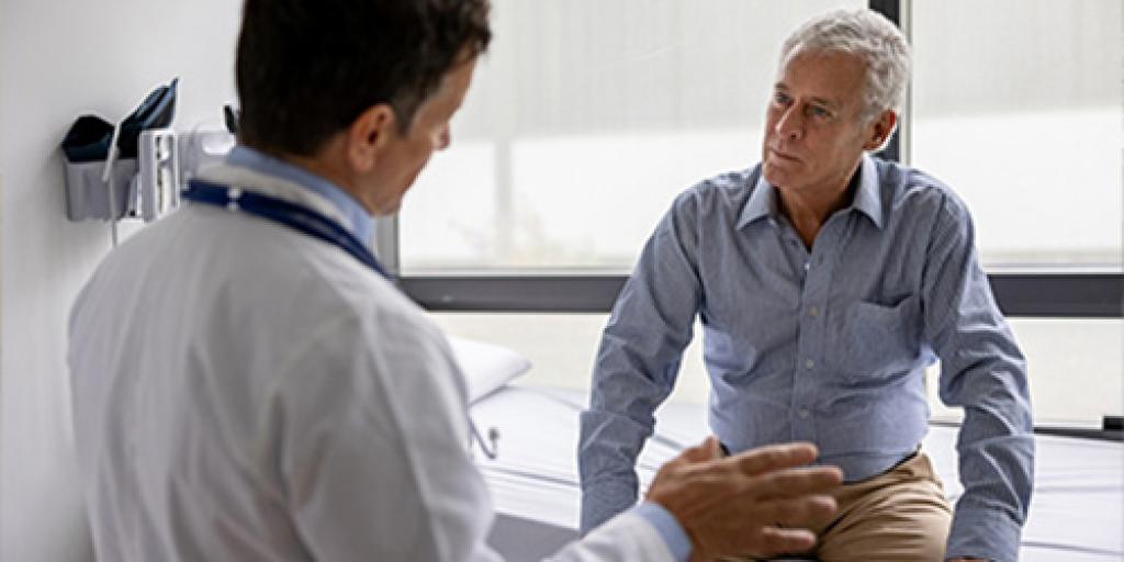 Doctor in white lab coat speaking with elderly man in exam room.
