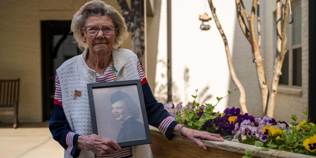 Margaret Brewer, U.S. Navy Veteran, poses in a flower garden at Somerby Baxter Village, in Fort Mill, SC, holding a photo of her in uniform. 