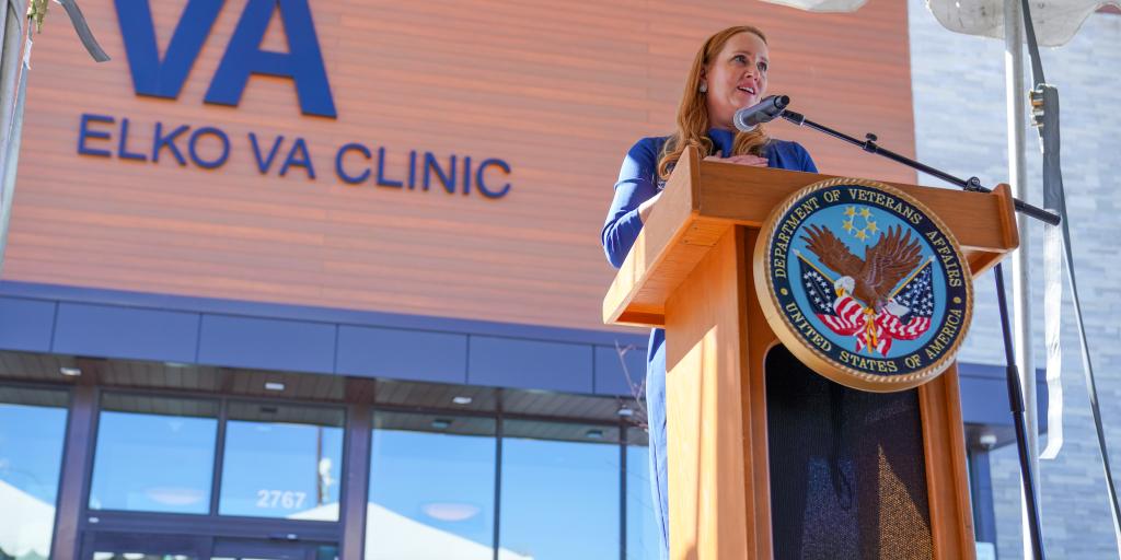 Woman at podium with microphone in front of Elko VA Clinic sign.