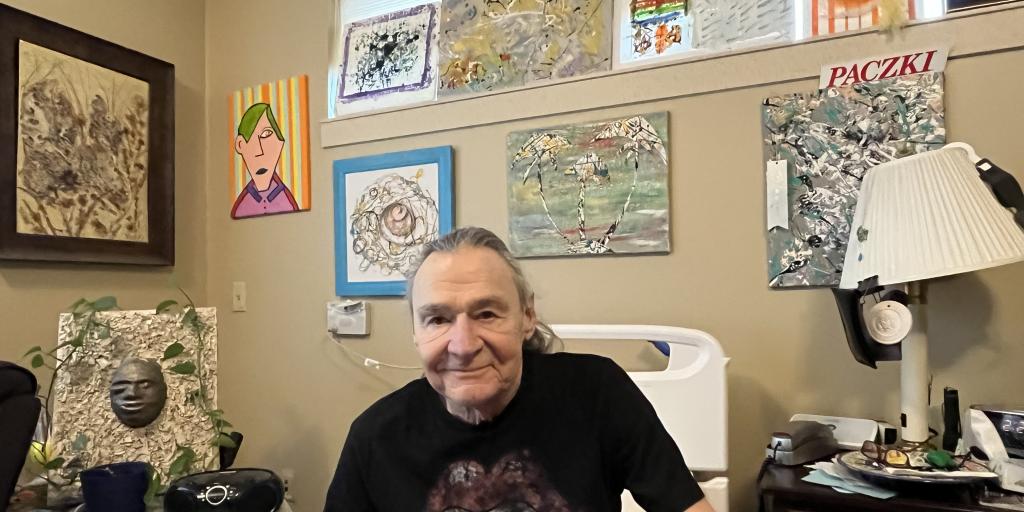 A smiling man in a black t-shirt with artwork on it, sitting on a red bed in a room with colorful framed art pieces.