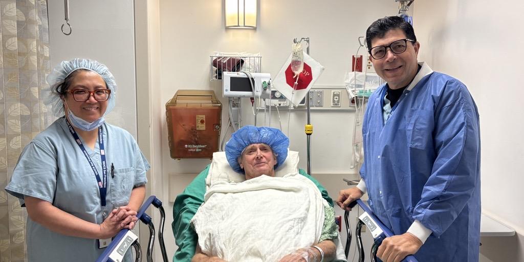 A patient in a hospital bed with two medical professionals prior to a procedure posing for a photo, all smiling.