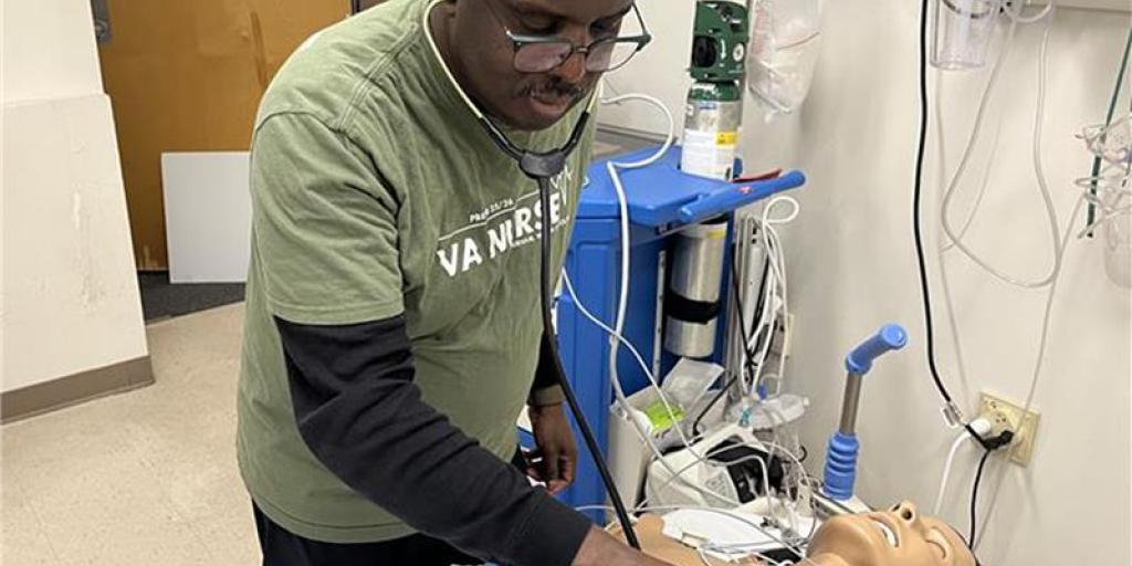 Man in green shirt and black pants using stethoscope on CPR mannequin in hospital room.