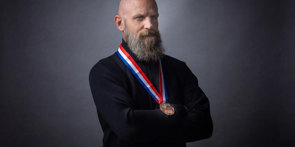 Bald man with a beard wearing a black shirt and a medal.