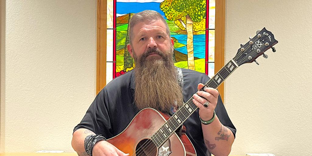 Bearded man with tattoos holding acoustic guitar in front of stained glass window.